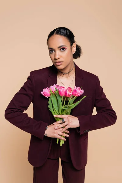 Fashionable african american woman in burgundy suit posing with pink tulips isolated on beige — Stock Photo