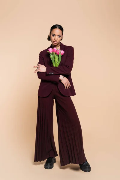 Повна довжина добре одягненої афроамериканки в бордовий костюм позує з рожевими тюльпанами на бежевому — стокове фото
