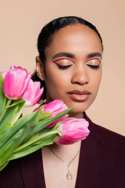 Encantadora mujer afroamericana con los ojos cerrados posando con tulipanes rosados aislados en beige - foto de stock