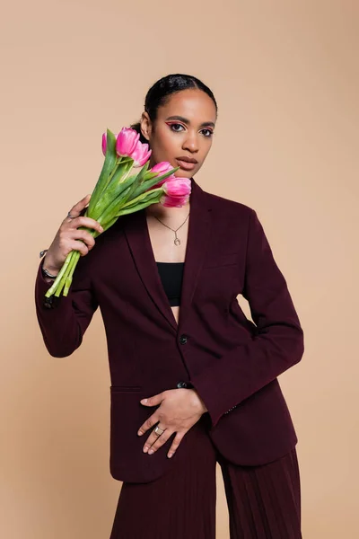 Well dressed african american woman in burgundy suit posing with tulips isolated on beige — Stock Photo