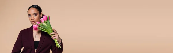 Stylisches afrikanisch-amerikanisches Model in weinrotem Blazer posiert mit rosa Tulpen isoliert auf beige, Banner — Stockfoto