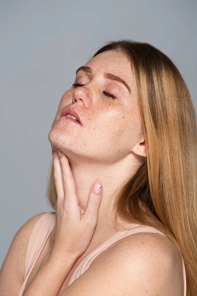 Junge Frau mit geschlossenen Augen berührt sommersprossige Haut isoliert auf grau — Stockfoto