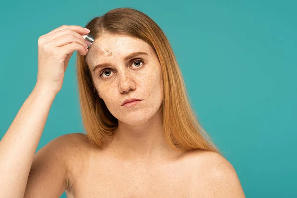 Woman with freckled skin applying serum on forehead with dropper isolated on turquoise — Stock Photo