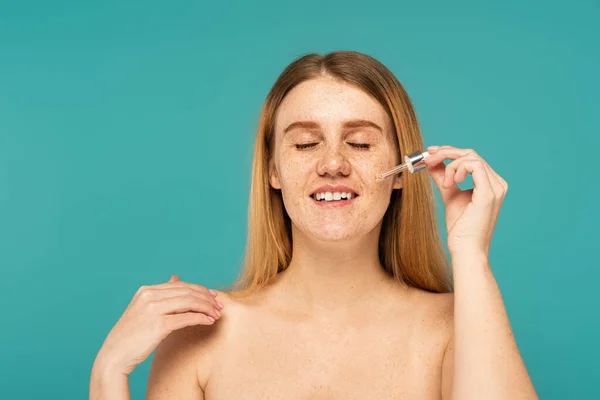 Smiling freckled woman applying serum on face isolated on turquoise — Stock Photo