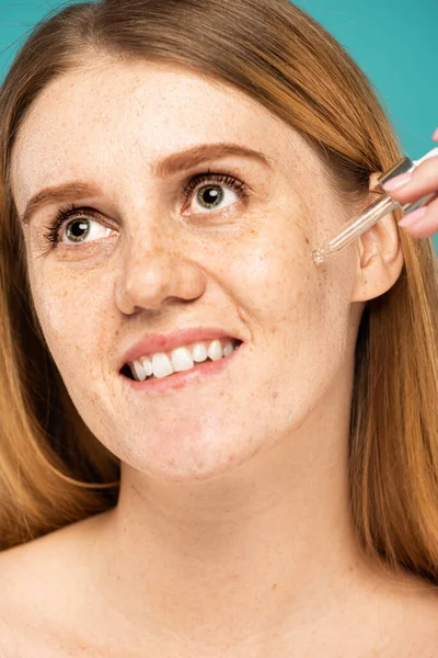 Retrato de mujer pecosa sonriente aplicando suero cosmético aislado en turquesa - foto de stock