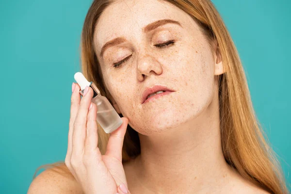 Young freckled model with closed eyes holding cosmetic serum isolated on turquoise — Stock Photo