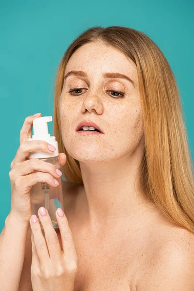 Jeune rousse femme aux taches de rousseur tenant bouteille avec nettoyant mousse près du visage isolé sur turquoise — Photo de stock