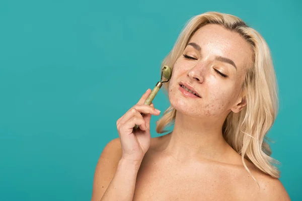 Blonde woman with acne smiling while using jade roller isolated on turquoise — Stock Photo