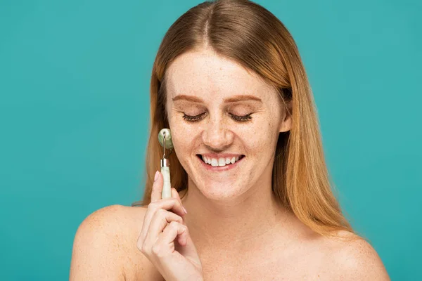 Happy redhead woman with freckles using jade roller isolated on turquoise — Stock Photo