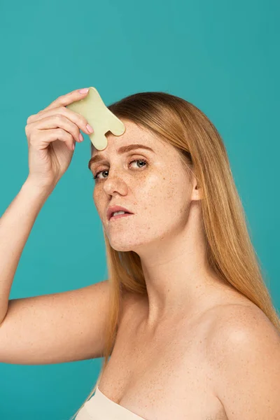 Young and redhead woman with freckles using face scrapper isolated on turquoise — Stock Photo