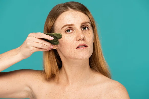 Jeune et rousse femme sans maquillage en utilisant gua sha visage scrapper isolé sur turquoise — Photo de stock