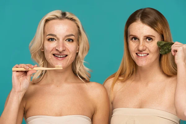 Femme blonde gaie brossant les dents près du modèle rousse en utilisant le grattoir visage isolé sur turquoise — Photo de stock