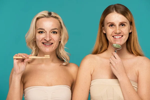 Cheerful blonde woman brushing teeth near redhead model using jade roller isolated on turquoise — Stock Photo