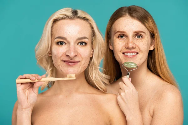 Glücklich blonde Frau Zähne putzen in der Nähe fröhlich Modell mit Jade-Rolle isoliert auf Türkis — Stockfoto