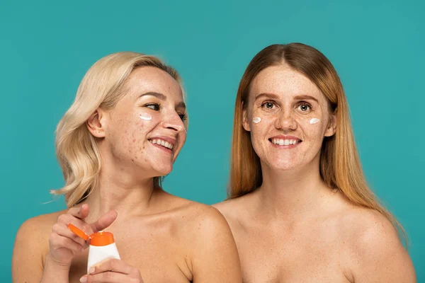 Mulheres jovens felizes com diferentes condições de pele aplicando creme em rostos isolados em turquesa — Fotografia de Stock