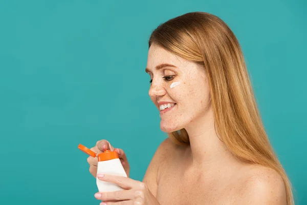 Cheerful redhead woman with freckles holding tube with moisturizing cream isolated on turquoise — Stock Photo