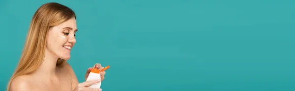 Femme rousse gaie avec des taches de rousseur tenant tube avec crème hydratante isolé sur turquoise, bannière — Photo de stock