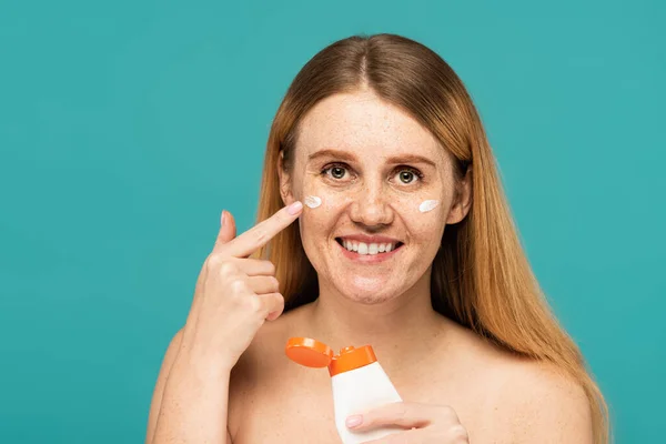 Mulher feliz com sardas segurando tubo e aplicando creme cosmético isolado em turquesa — Fotografia de Stock
