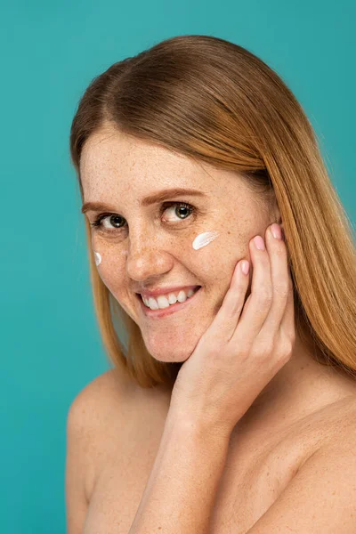 Cheerful woman with freckles and cosmetic cream on face isolated on turquoise — Stock Photo