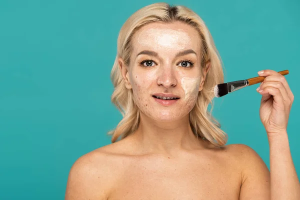 Blondine mit Akne hält Kosmetikpinsel in der Hand und trägt Tonmaske isoliert auf Türkis auf — Stockfoto