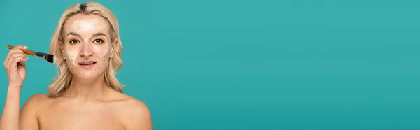 Femme blonde avec acné tenant pinceau cosmétique et appliquant masque d'argile isolé sur turquoise, bannière — Photo de stock