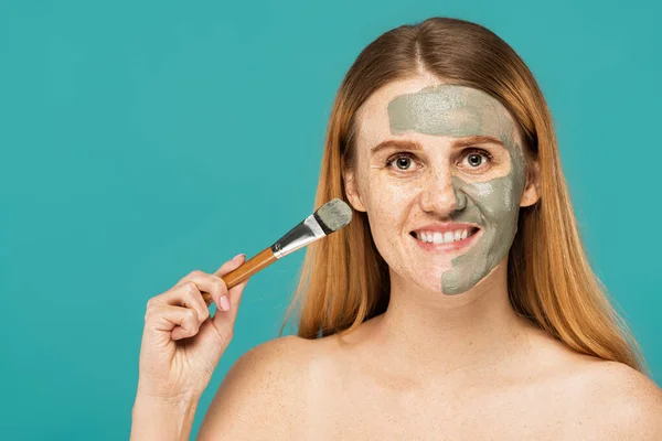 Весела жінка з рудим волоссям тримає косметичну щітку при нанесенні глиняної маски на половину обличчя ізольовано на бірюзовому — стокове фото
