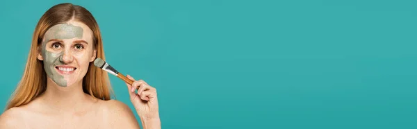 Femme gaie aux cheveux roux tenant pinceau cosmétique tout en appliquant un masque d'argile sur le visage isolé sur turquoise, bannière — Photo de stock