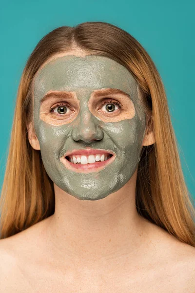 Cheerful redhead woman with clay mask on face smiling isolated on turquoise — Stock Photo