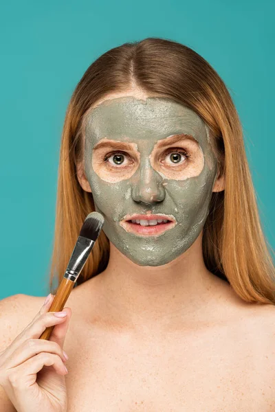 Portrait de jolie jeune femme aux cheveux roux appliquant un masque d'argile sur le visage isolé sur turquoise — Photo de stock