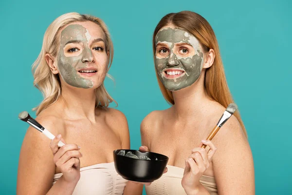 Blonde and redhead women applying clay mask on faces while holding cosmetic brushes isolated on turquoise — Stock Photo
