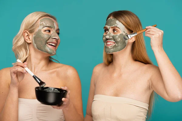 Mujeres alegres aplicando máscara de arcilla en las caras mientras sostiene cepillos cosméticos aislados en turquesa - foto de stock