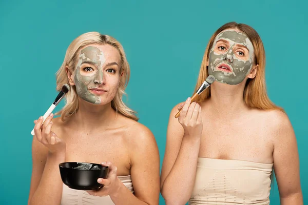 Femmes blondes et rousses appliquant un masque d'argile sur les visages avec des pinceaux cosmétiques isolés sur turquoise — Photo de stock