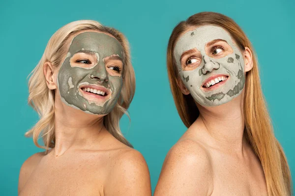 Ruiva alegre e mulheres loiras com máscara de barro em rostos posando isolado em turquesa — Fotografia de Stock