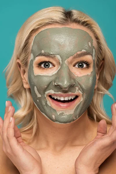 Excited blonde woman with clay mask on face looking at camera isolated on turquoise — Stock Photo