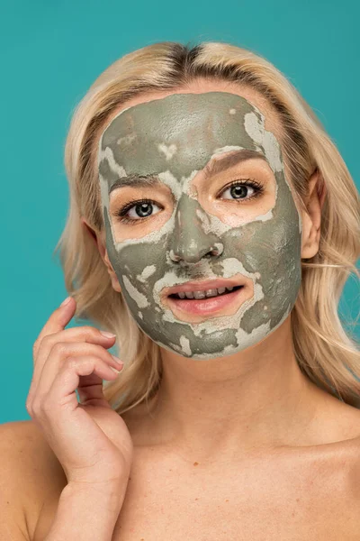 Portrait of cheerful blonde woman with clay mask on face looking at camera isolated on turquoise — Stock Photo