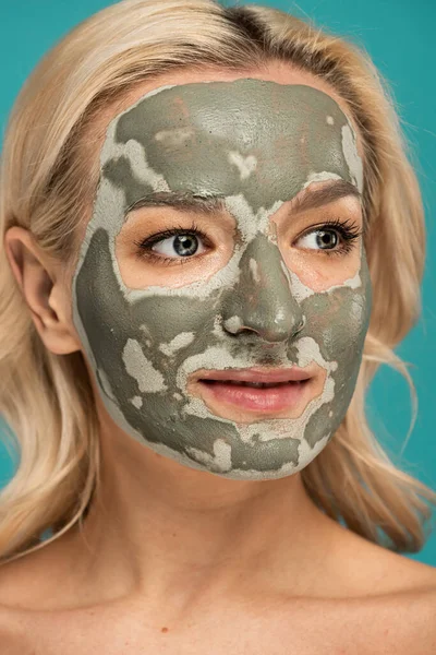Retrato de mujer rubia con máscara de arcilla en la cara mirando hacia otro lado aislado en turquesa - foto de stock