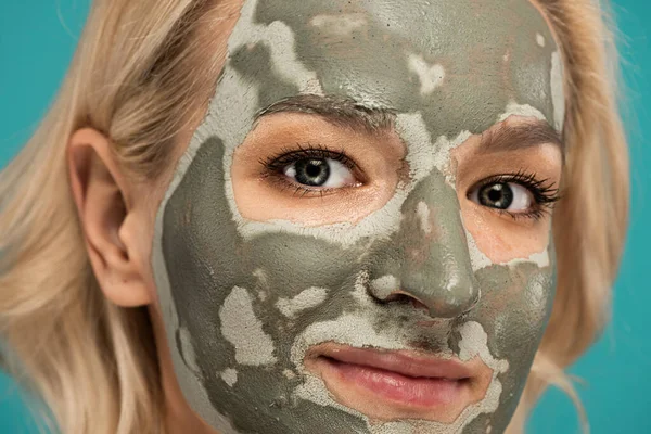 Gros plan de femme blonde souriante avec masque en argile sur le visage en regardant la caméra isolée sur turquoise — Photo de stock
