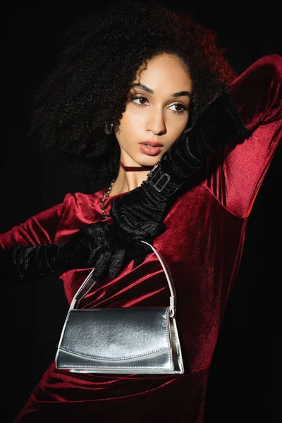 Modelo afroamericano de moda en vestido de terciopelo y guantes con bolso de plata aislado en negro - foto de stock