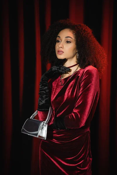 Elegante donna afroamericana in abito e guanti in possesso di borsa su sfondo drappeggio bordeaux — Foto stock