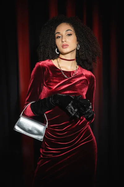 Elegante mujer afroamericana joven en vestido posando con bolso de mano sobre fondo borgoña - foto de stock