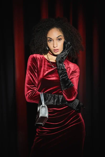 Femme afro-américaine élégante en gants de velours et robe posant avec sac à main sur fond de draperie bordeaux — Photo de stock