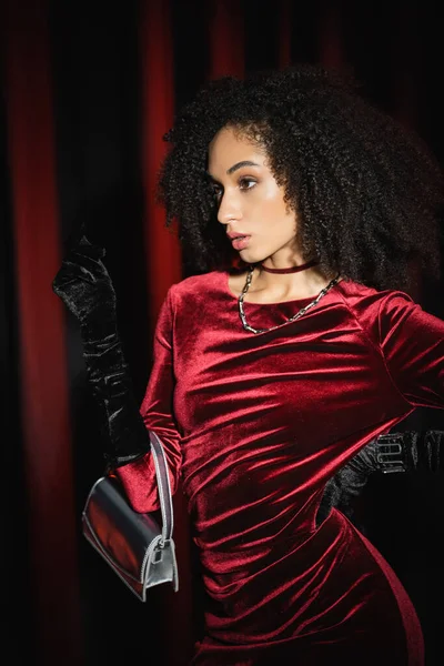 Porträt einer Afroamerikanerin in elegantem Kleid, die mit Handtasche auf weinrotem Vorhang posiert — Stockfoto