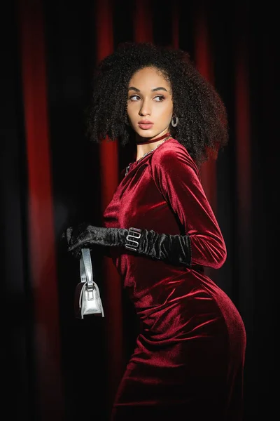 Femme afro-américaine élégante en robe tenant sac à main sur fond de draperie bordeaux — Photo de stock