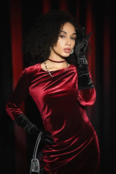 Jolie modèle afro-américain posant en robe de velours et tenant un sac à main près de draperies bordeaux — Photo de stock