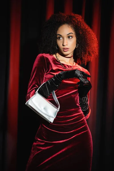 Mujer afroamericana en vestido de terciopelo sosteniendo el bolso y mirando hacia otro lado cerca de cortinas borgoña - foto de stock