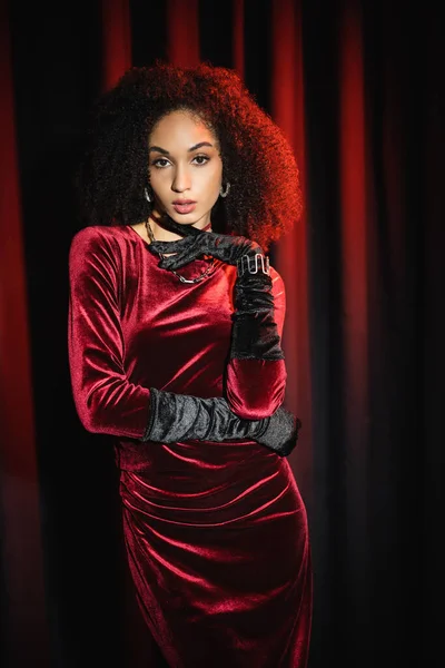 Modelo afroamericano en vestido de terciopelo y guantes mirando a la cámara cerca de cortinas rojas - foto de stock