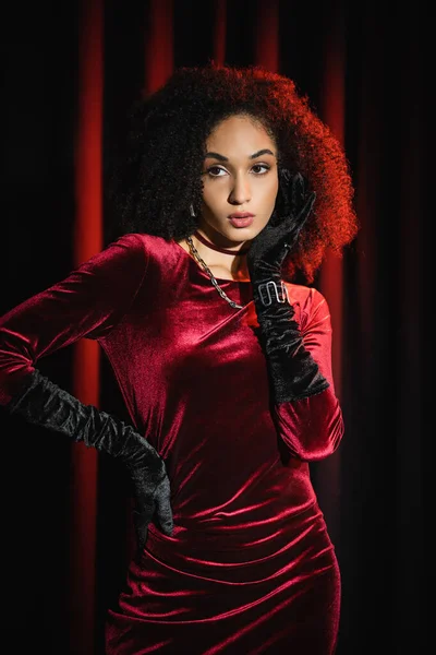 Modische afrikanisch-amerikanische Frau in Samtkleid und Handschuhen, die das Gesicht in der Nähe roter Vorhänge berühren — Stockfoto