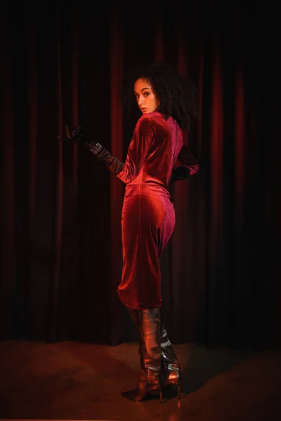 Трендовая африканская женщина в сапогах на коленях и платье смотрит в камеру рядом с красной драпировкой — стоковое фото
