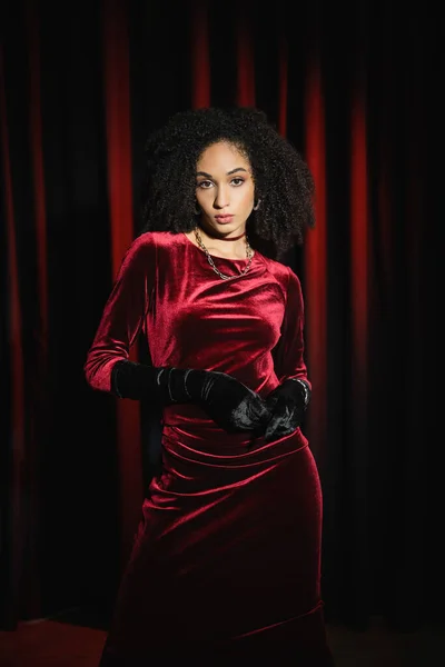 Elegante modelo afroamericano en vestido de terciopelo y guantes mirando a la cámara cerca de cortinas rojas - foto de stock