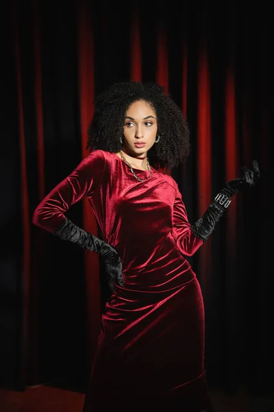 Elegante mujer afroamericana en guantes y vestido posando cerca de cortinas rojas - foto de stock
